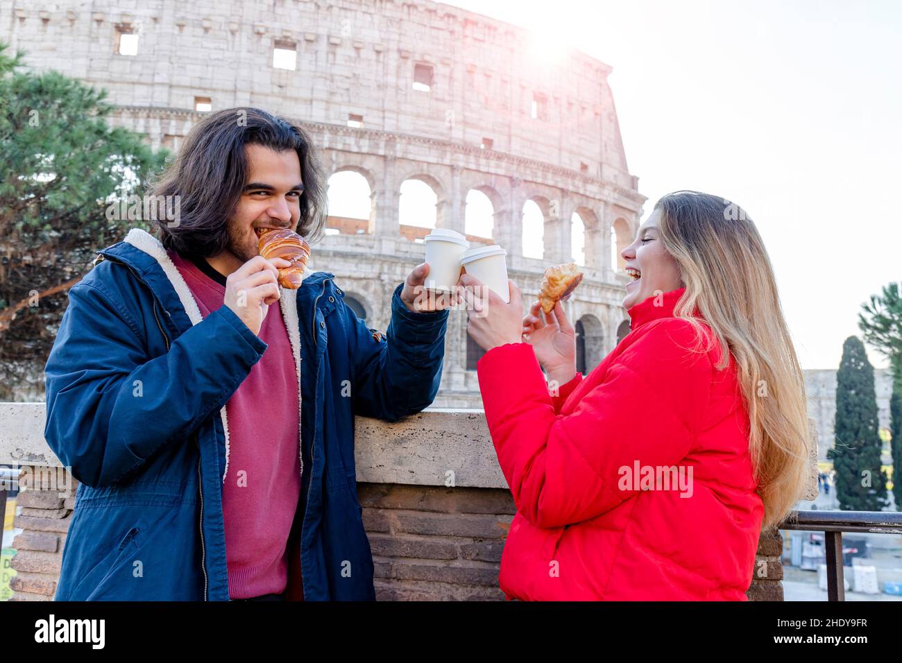 Young couple traveling to Rome. The couple is eating a croissant and drinking a coffee in front of the Colosseum. Stock Photo