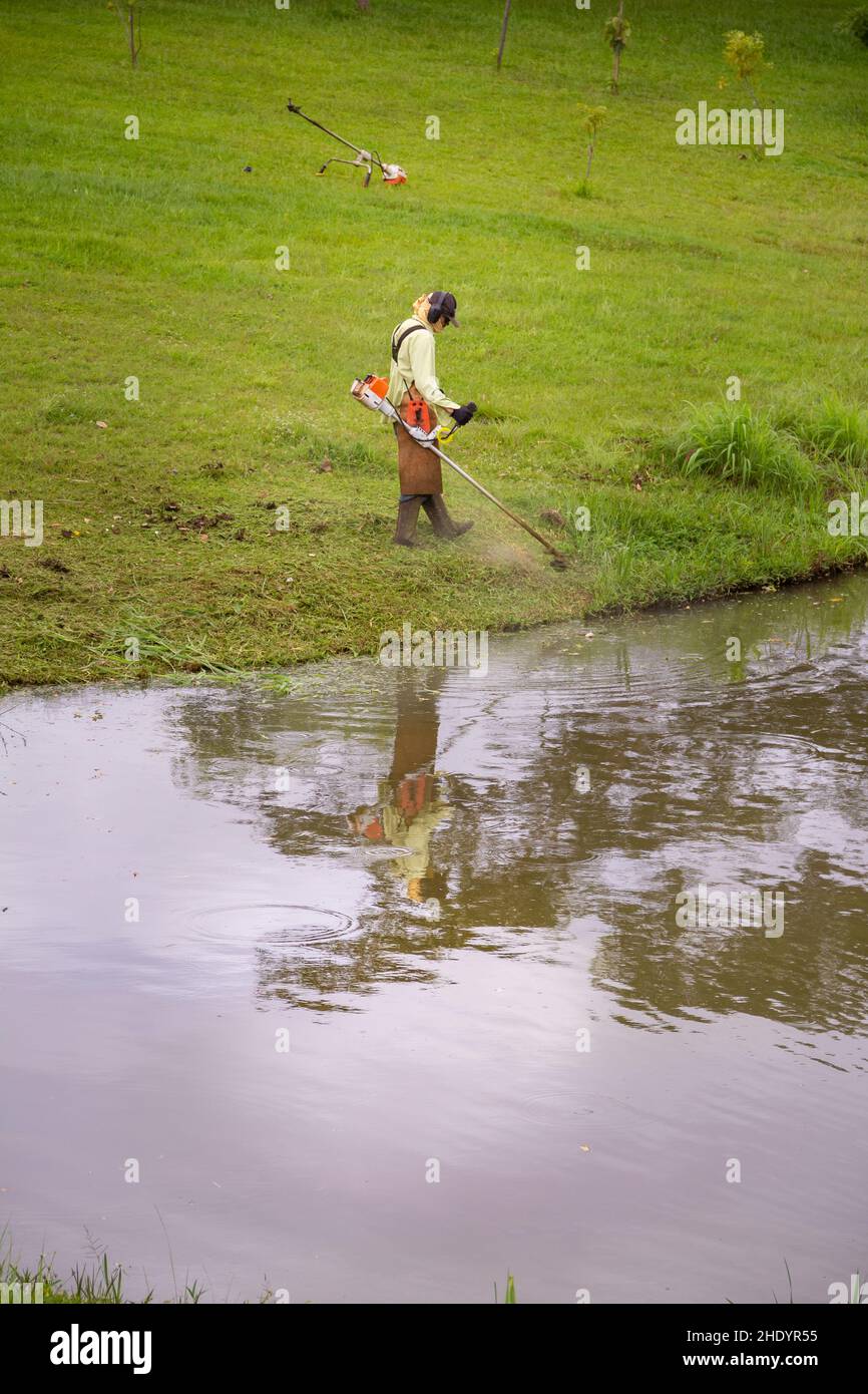 Goiânia, Goias, Brazil – January 05, 2022: A worker who cuts grass at the edge of a lake using appropriate equipment. Stock Photo