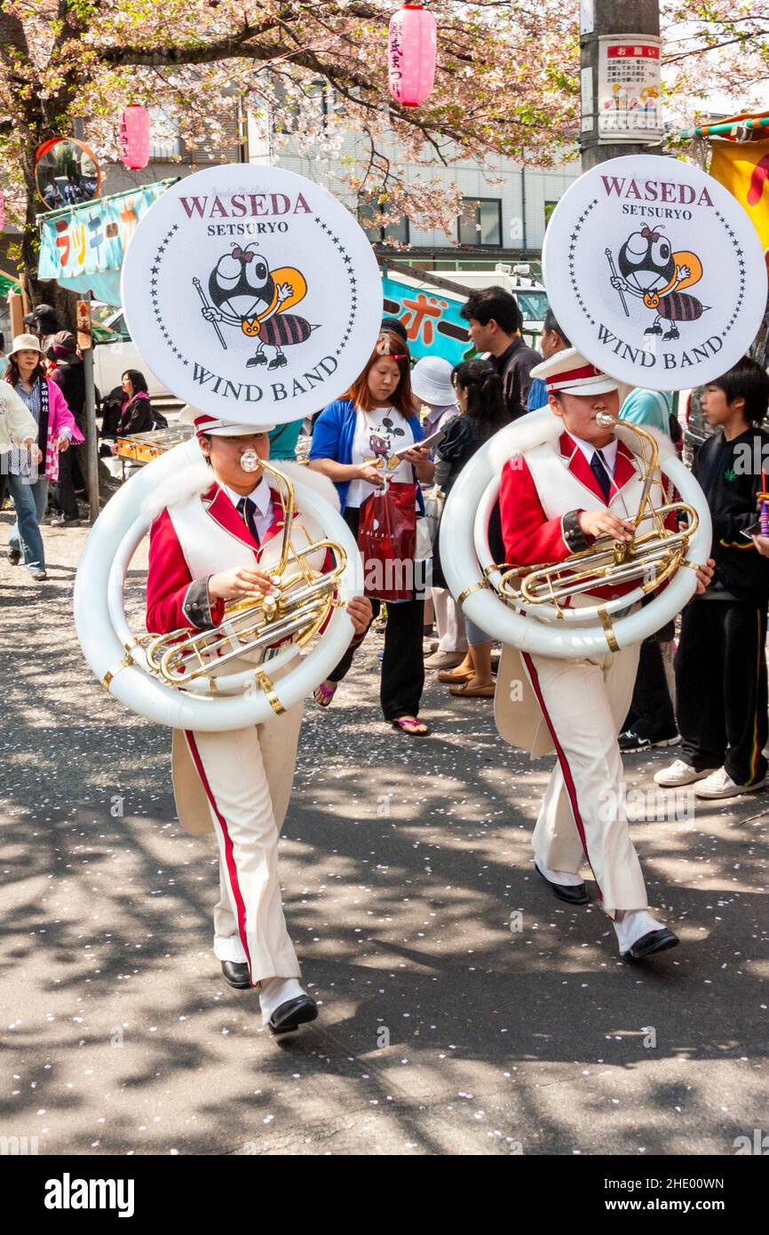 2 Japanese members of the Waseda Band in pink and white military style uniform and playing French horn while marching in parade under cherry blossoms. Stock Photo