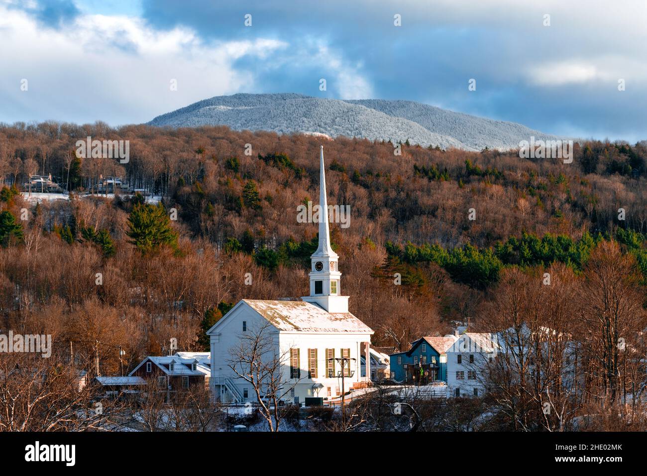 Small village surrounded by pine and maple tree forest with snow covered mountains in background. Stock Photo