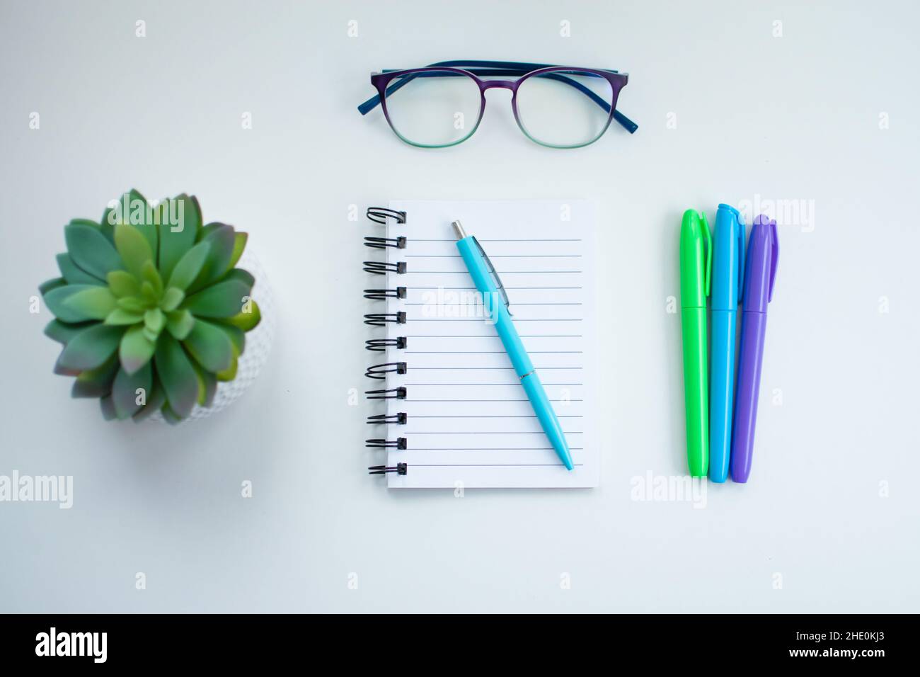 High angle of a white desk with colorful writing instruments on it and a potted green plant, glasses and an empty notebook Stock Photo