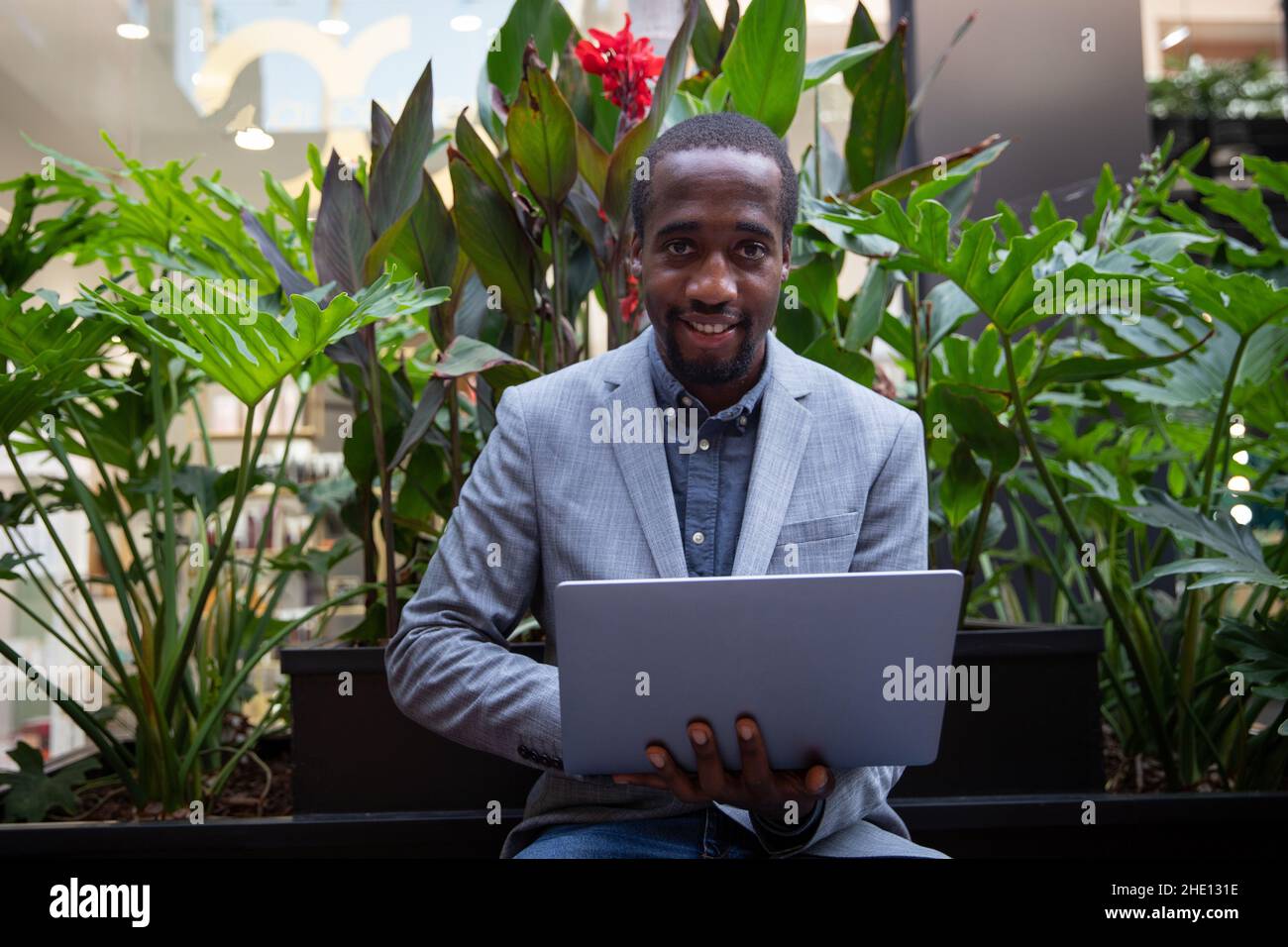 African businessman sitting on a bench works with his laptop. Successful person with confidant look smiles. Businessman works outdoors Stock Photo