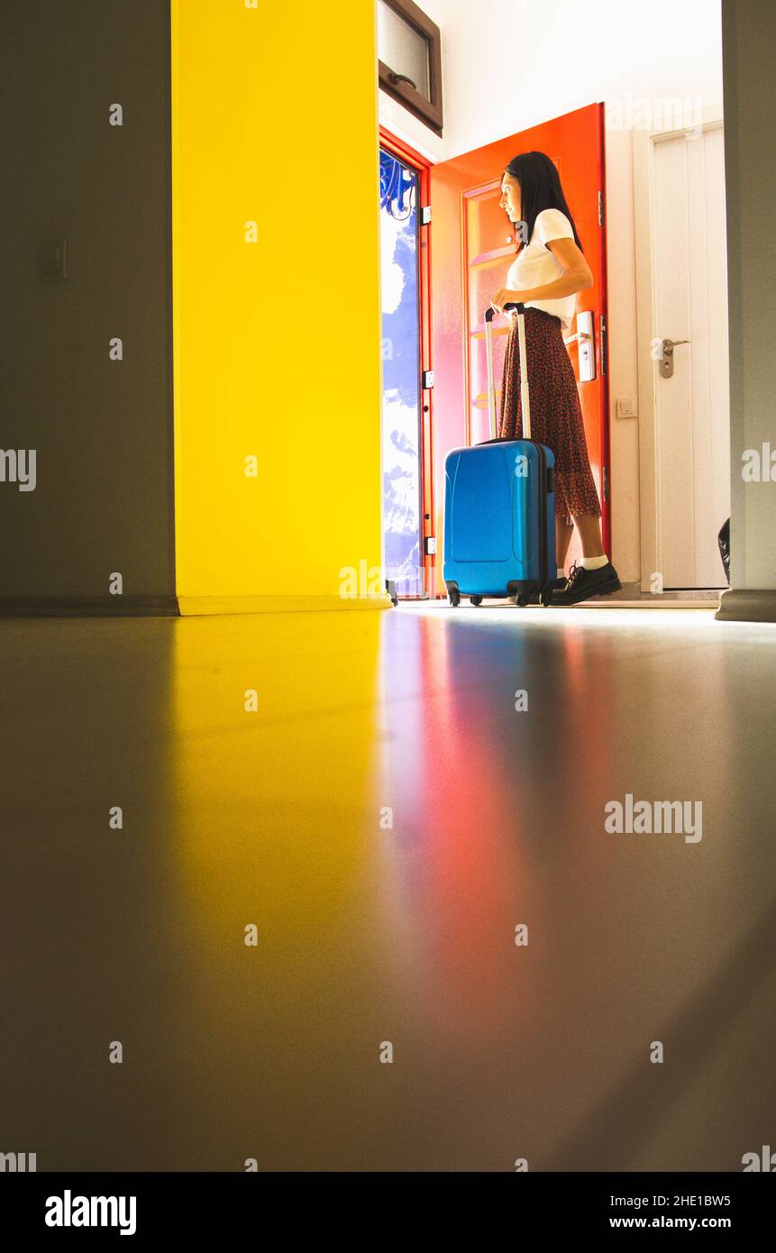 Young caucasian woman in casual clothes pushes blue luggage towards the exit of colorful apartment. Blank space background holiday image. Stock Photo