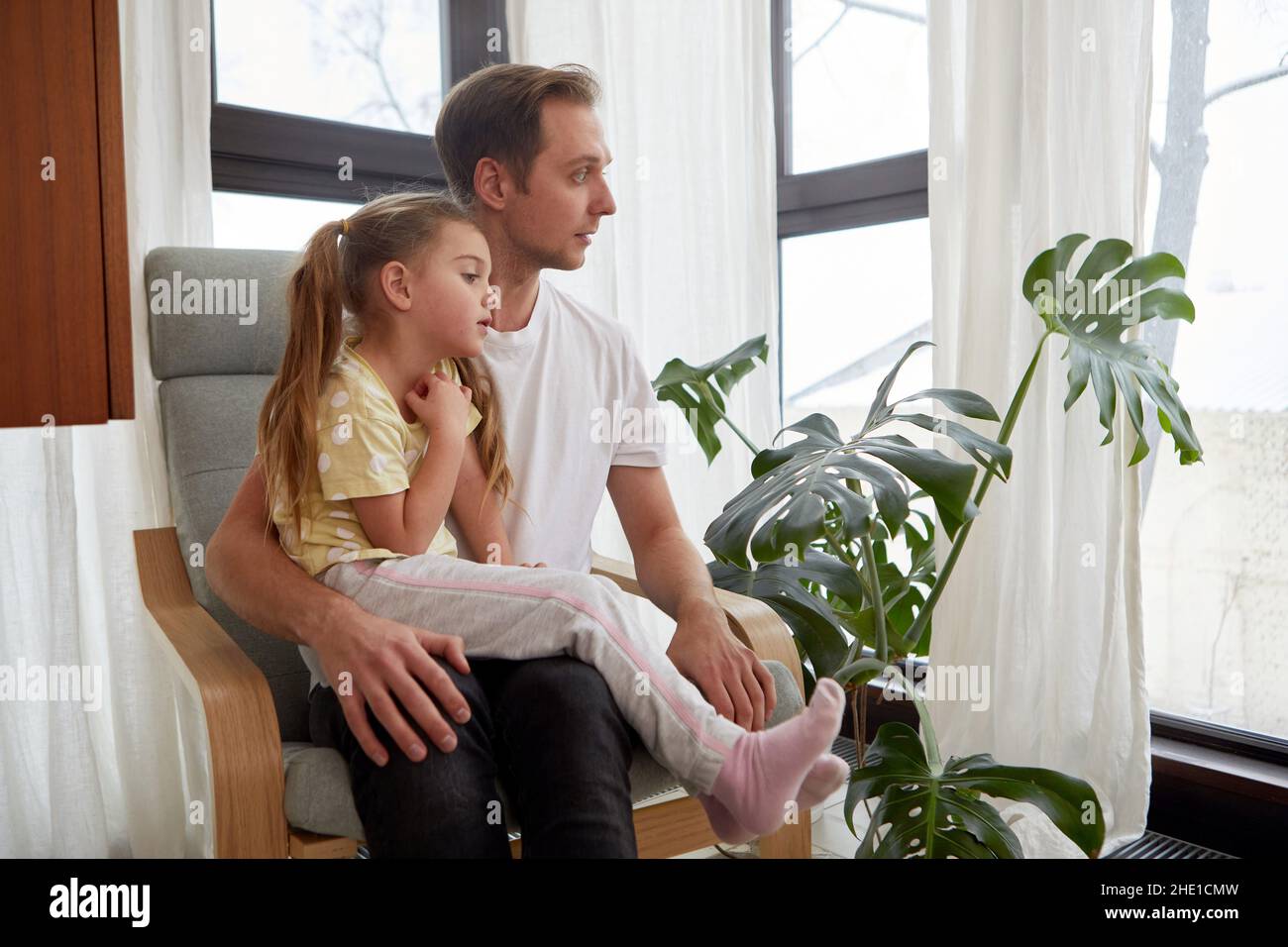 Dad and cute girl sitting on knees in armchair near potted plant and looking in window with attention Stock Photo