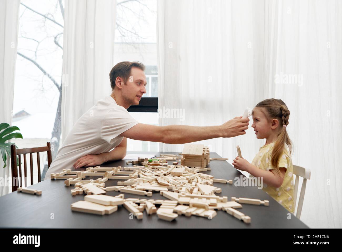 Side view of dad checking body temperature with infrared forehead thermometer of daughter sitting at table and playing with wooden blocks Stock Photo