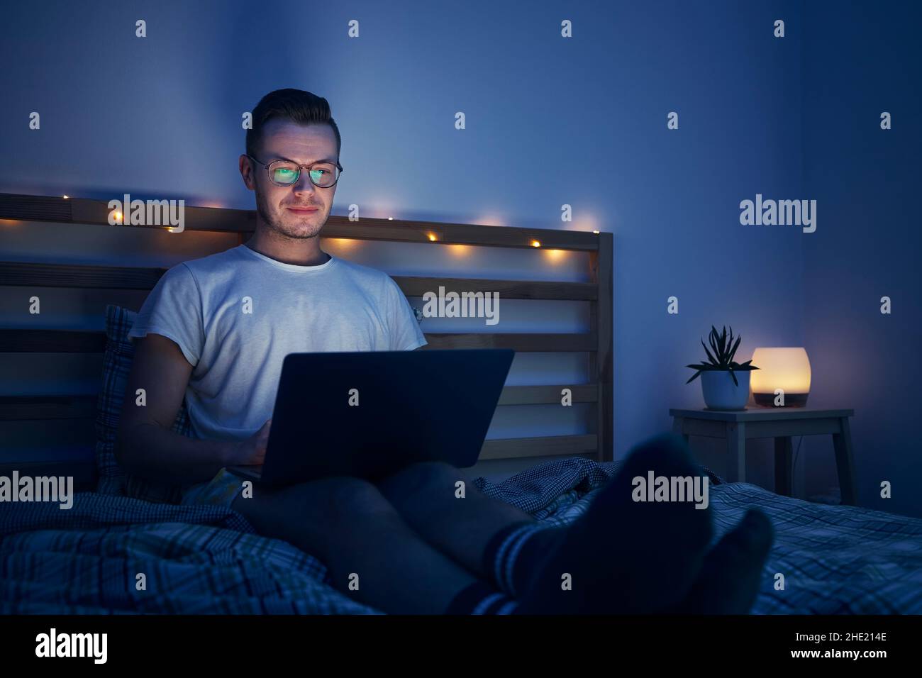 Man with eyeglasses sitting on bed in bedroom and using laptop. Face illuminated from computer display. Stock Photo