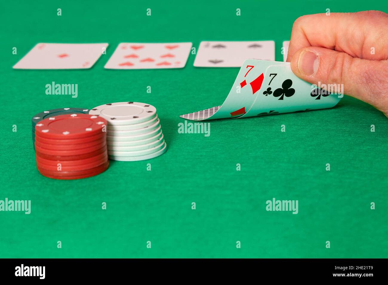On a green mat you can see a man's hand holding up two poker cards with his fingers, both cards are sevens. Next to them are stacked chips and in the Stock Photo