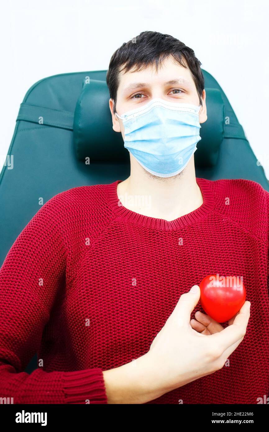 Young man in medical mask sits in chair and holds small heart in his hand. Grateful patient. Topic of health care and donation. Rescue of sick people Stock Photo