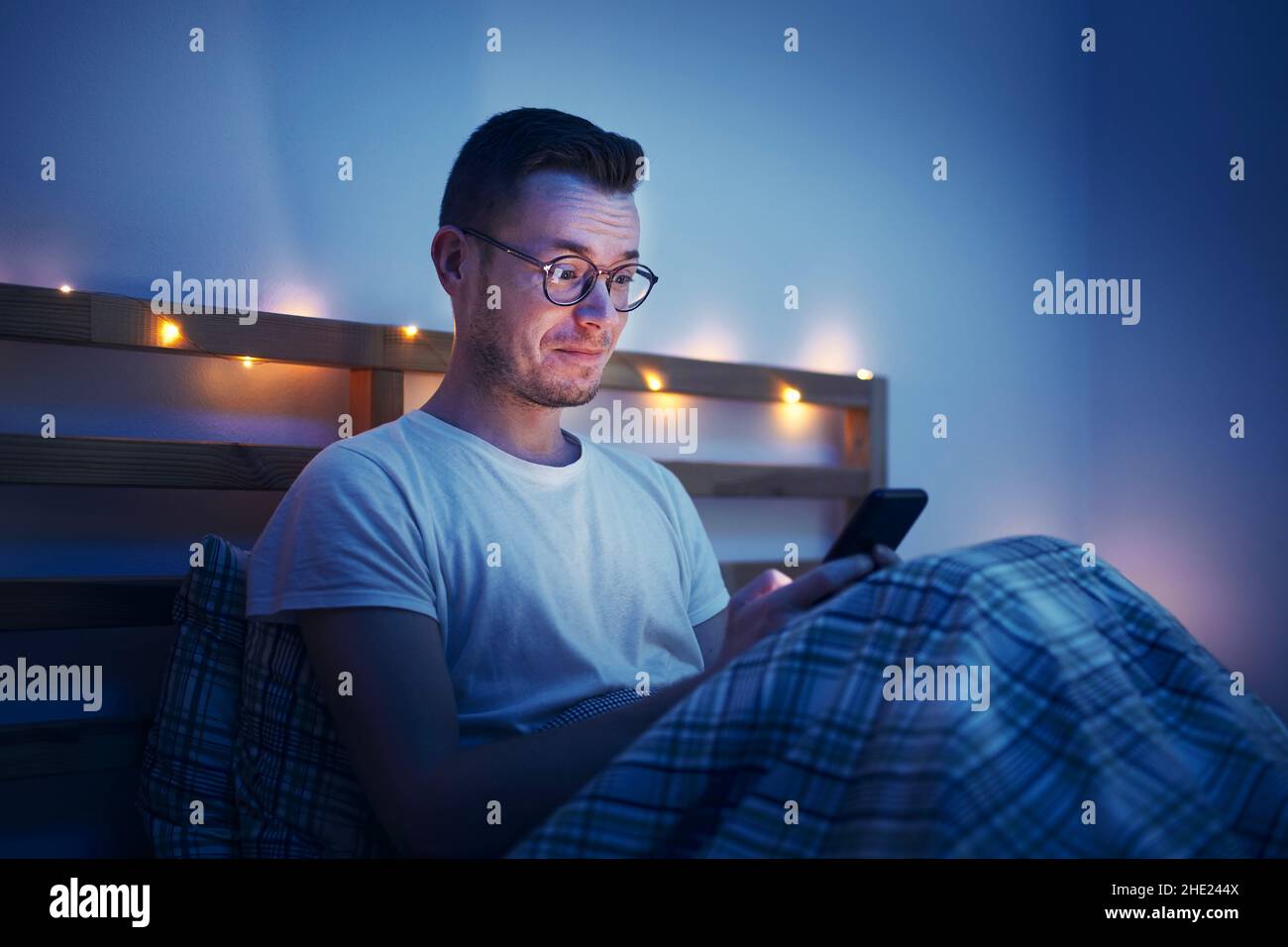 Man with eyeglasses sitting under duvet on bed in bedroom and using phone. Surprised face illuminated from computer display. Stock Photo