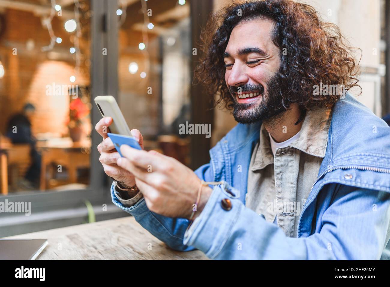 Gleeful Hispanic male in casual clothes with curly hair and beard smiling. He is entering credit card credentials into cellphone while sitting at cafeteria table and shopping online on street Stock Photo