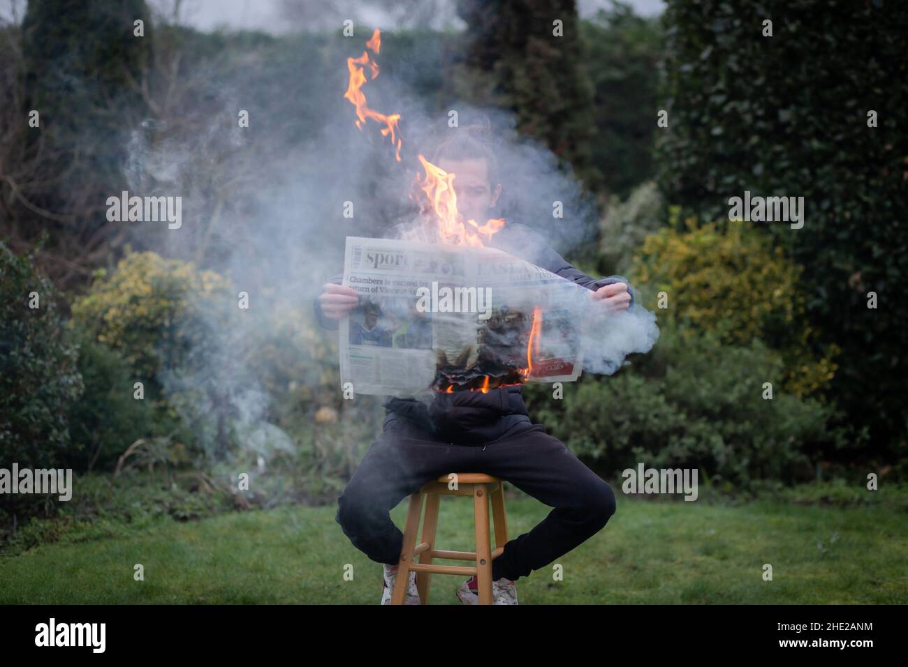 Woodbridge, Suffolk, UK January 01 2021: A portrait of a young man reading a burning newspaper while sitting outside. Hot news, breaking news concept Stock Photo