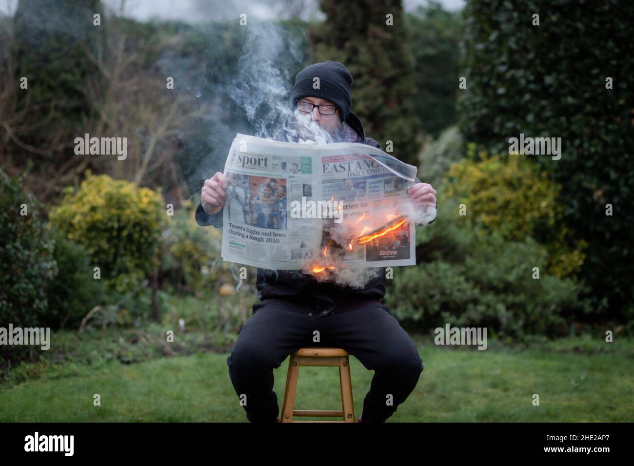 Woodbridge, Suffolk, UK January 01 2021: A portrait of a young man reading a burning newspaper while sitting outside. Hot news, breaking news concept Stock Photo