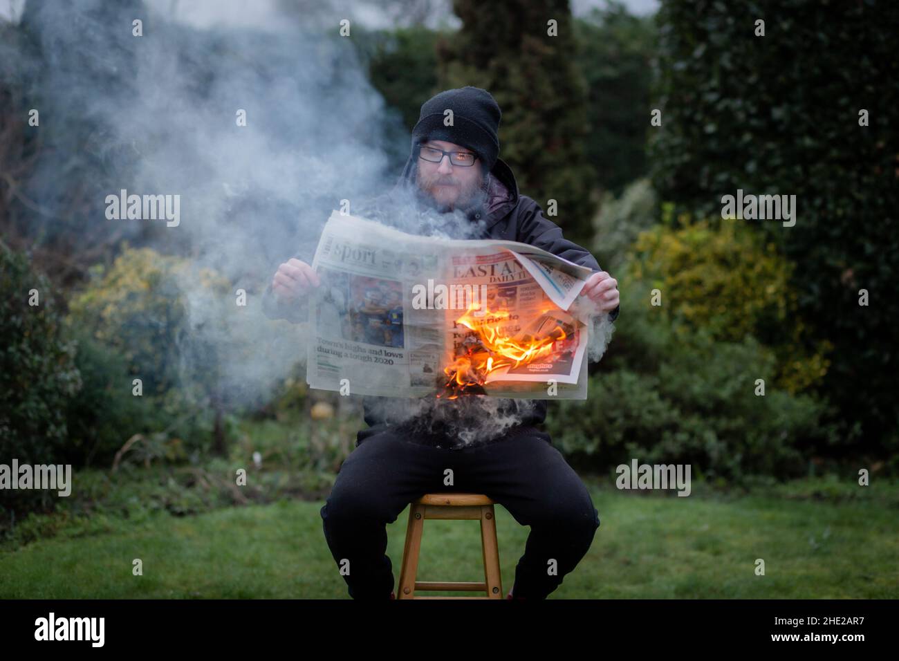 Woodbridge, Suffolk, UK January 01 2021: A portrait of a young man reading a burning newspaper while sitting outside. Hot news, breaking news concept Stock Photo