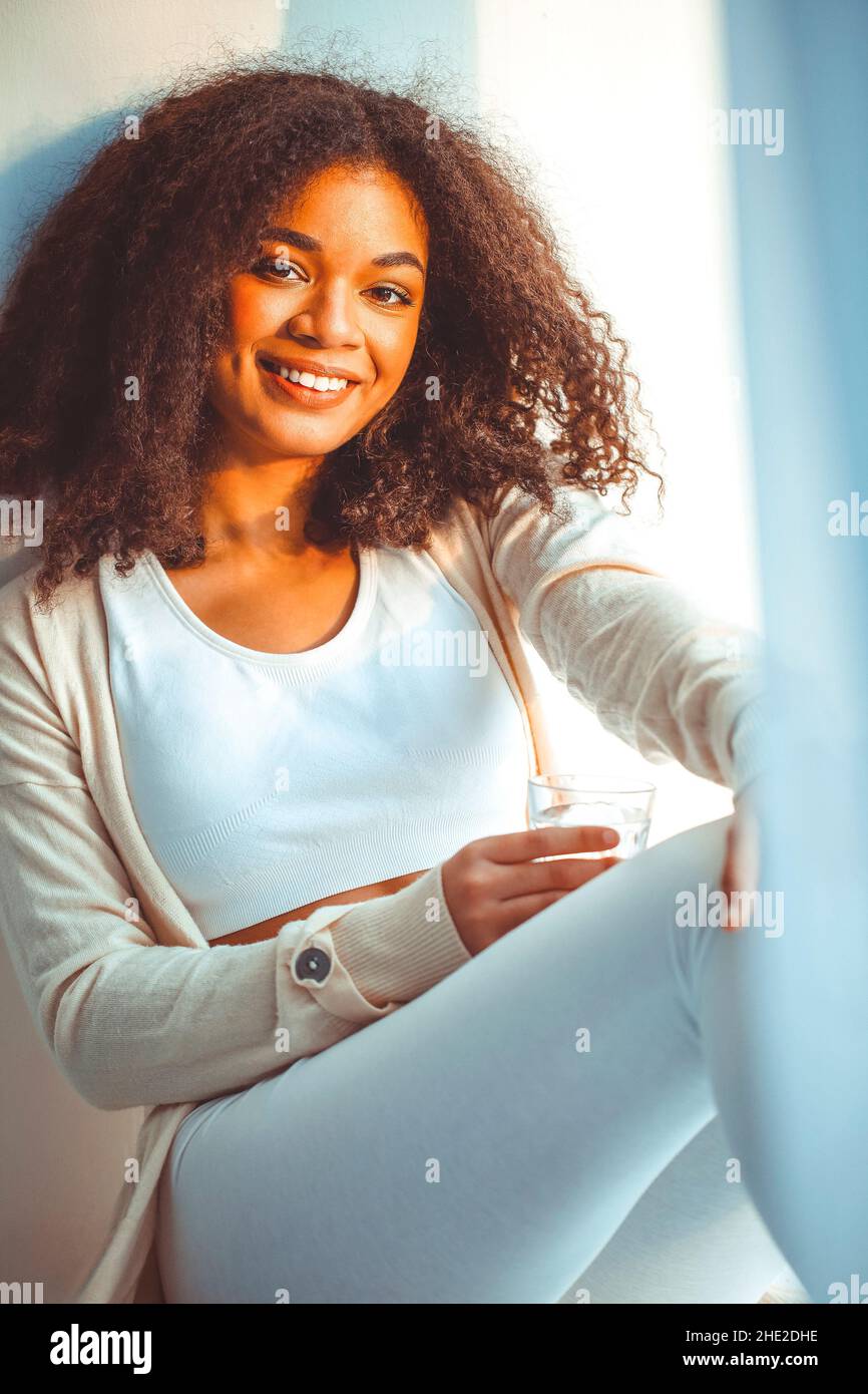 Young positive african american woman sitting on windowsill with glass ...
