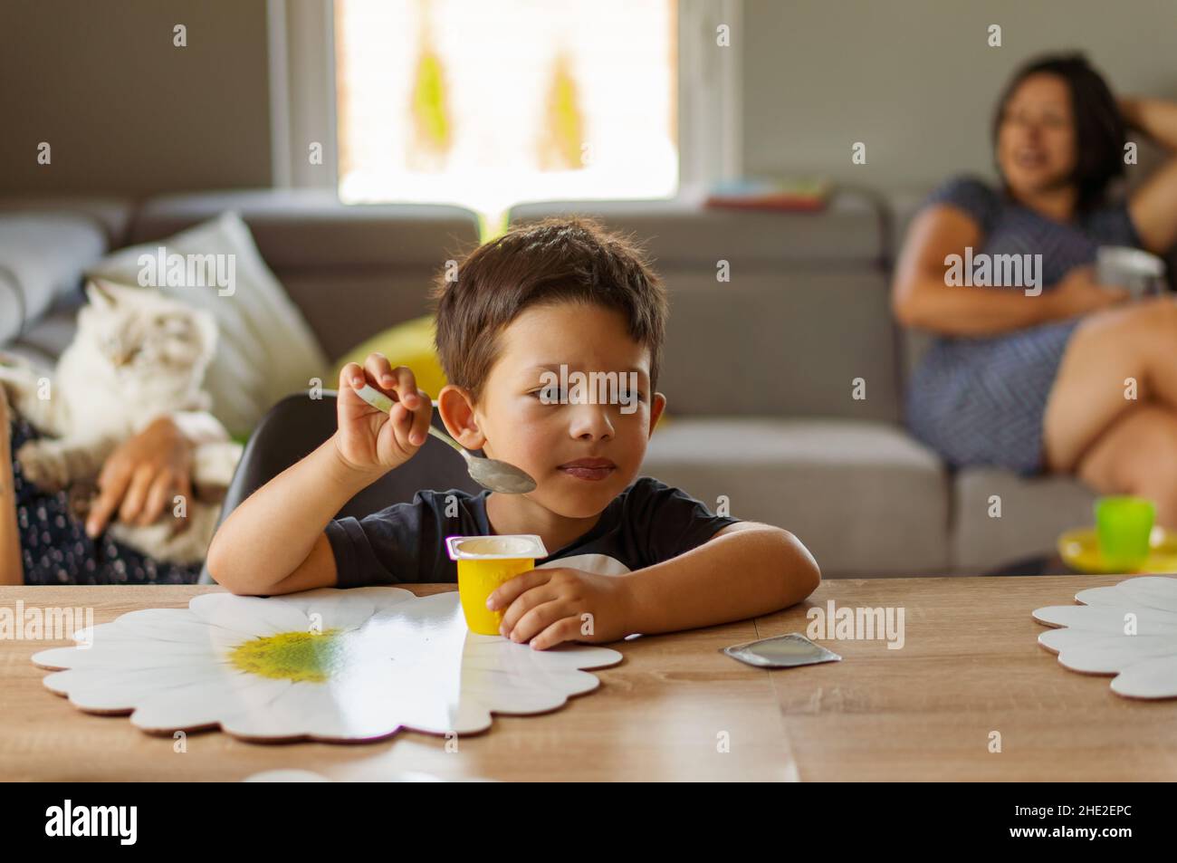Closeup shot of a young boy eating a yogurt with his mother sitting on the sofa on a background Stock Photo