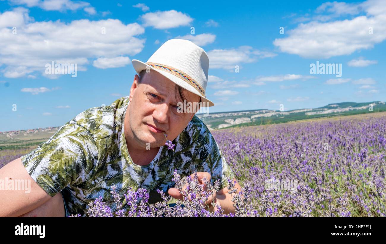 A smiling guy in a lilac field having a joyful face sits in a cap against a blue sky with a blue sky, and in a white cap Stock Photo