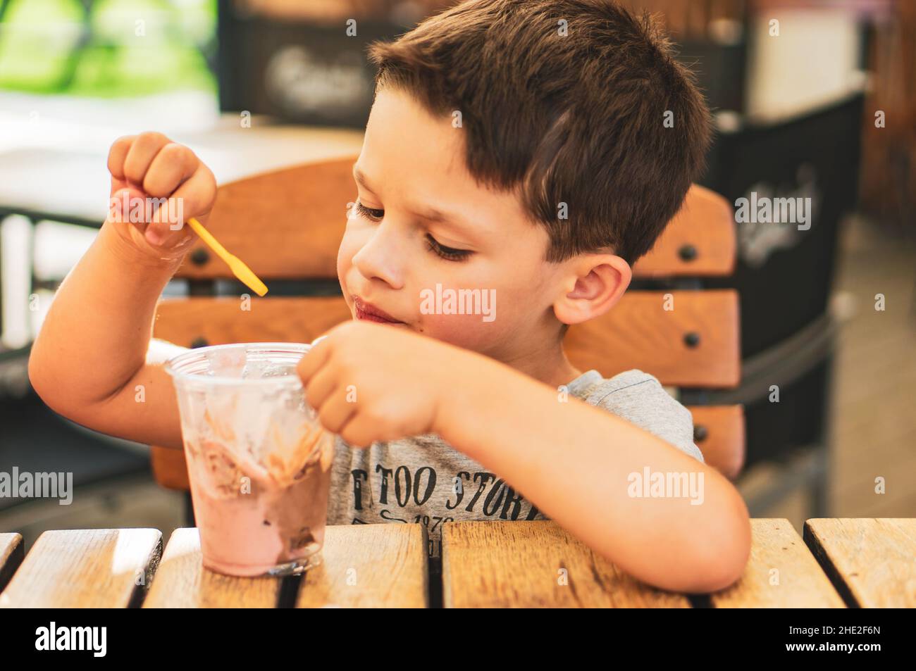 Closeup shot of a young boy eating ice cream from a plastic cup by a wooden table at a bar Stock Photo