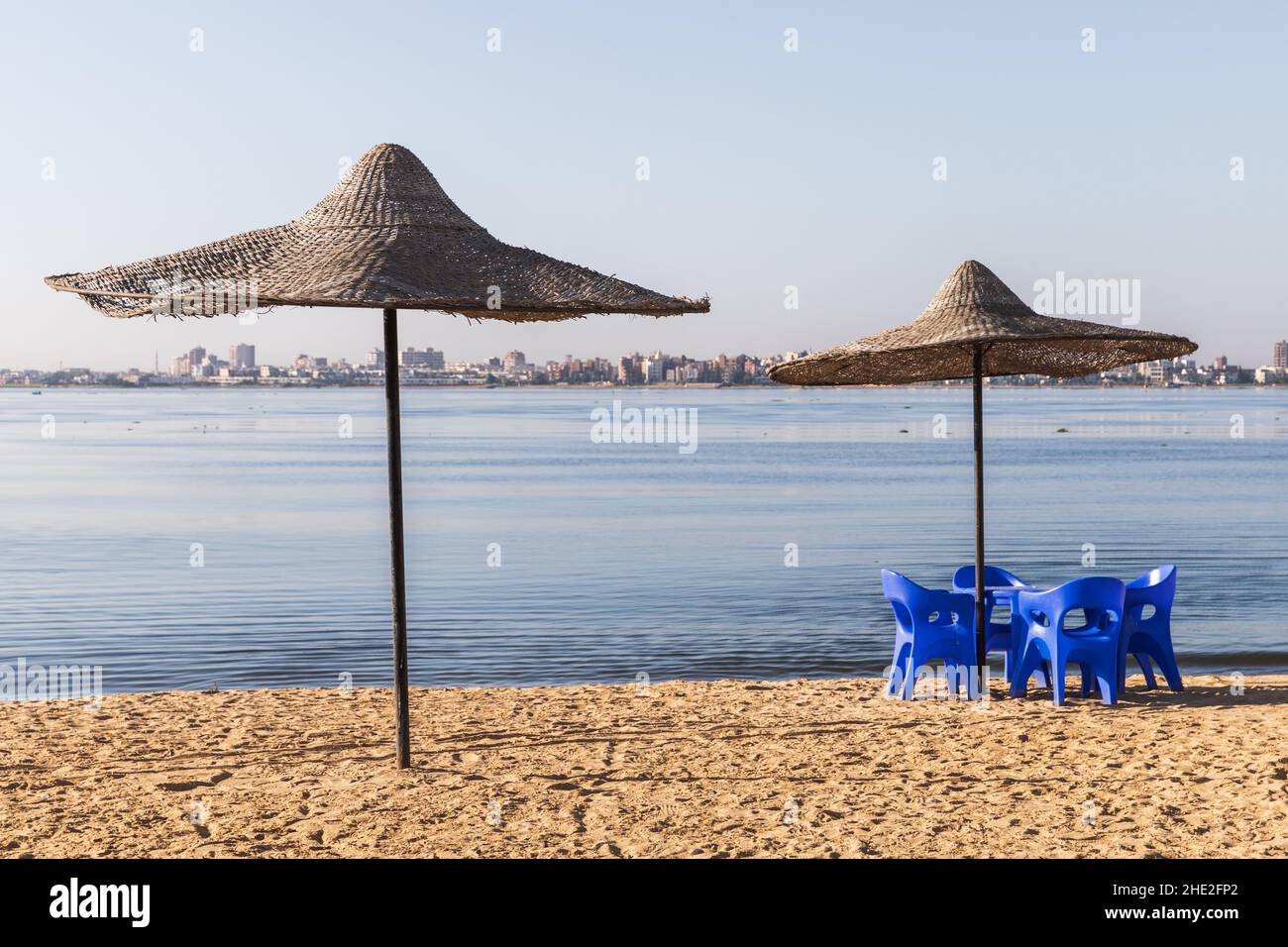 Straw umbrellas and blue plastic chairs are on the beach at Lake Timsah, one of the Bitter Lakes linked by the Suez Canal. Ismailia, Egypt Stock Photo