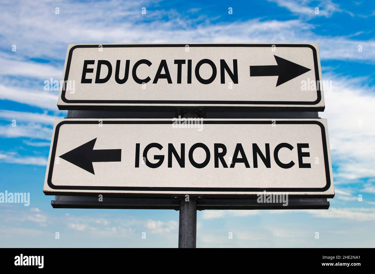 Education versus ignorance road sign with two arrows on blue and grey sky background. White two street sign with arrows on metal pole. Stock Photo