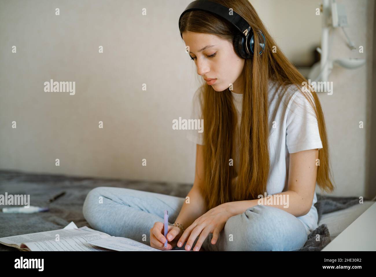 Young teenage girl in headphones does homework, sitting on bed Stock Photo
