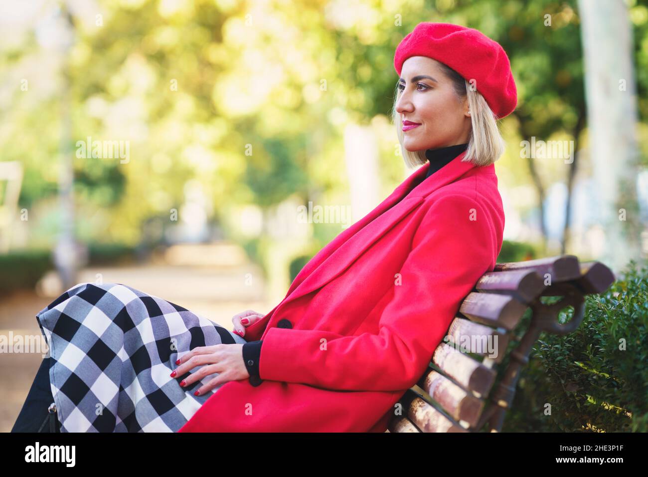 Middle-aged woman sitting on a bench in an urban park wearing red winter clothing. Stock Photo