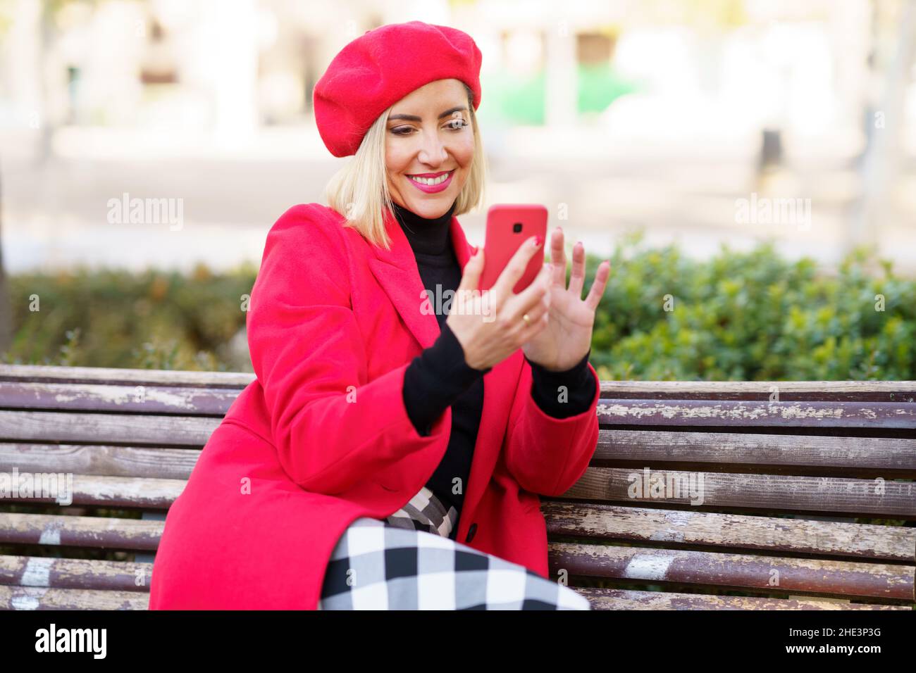 Middle-aged woman sitting on park bench using smartphone wearing red winter clothes. Stock Photo