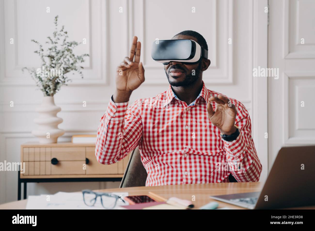 Focused Aframerican man in VR glasses enjoying augmented reality while sitting at workplace Stock Photo