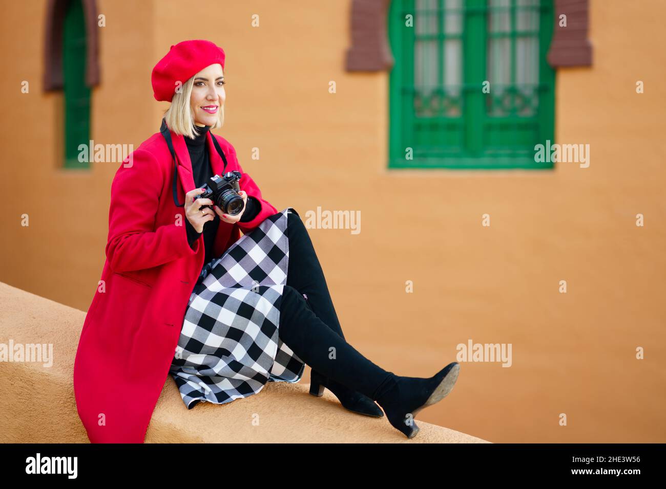 Woman wearing red winter clothes, taking pictures with an SLR camera sitting on a city wall. Stock Photo