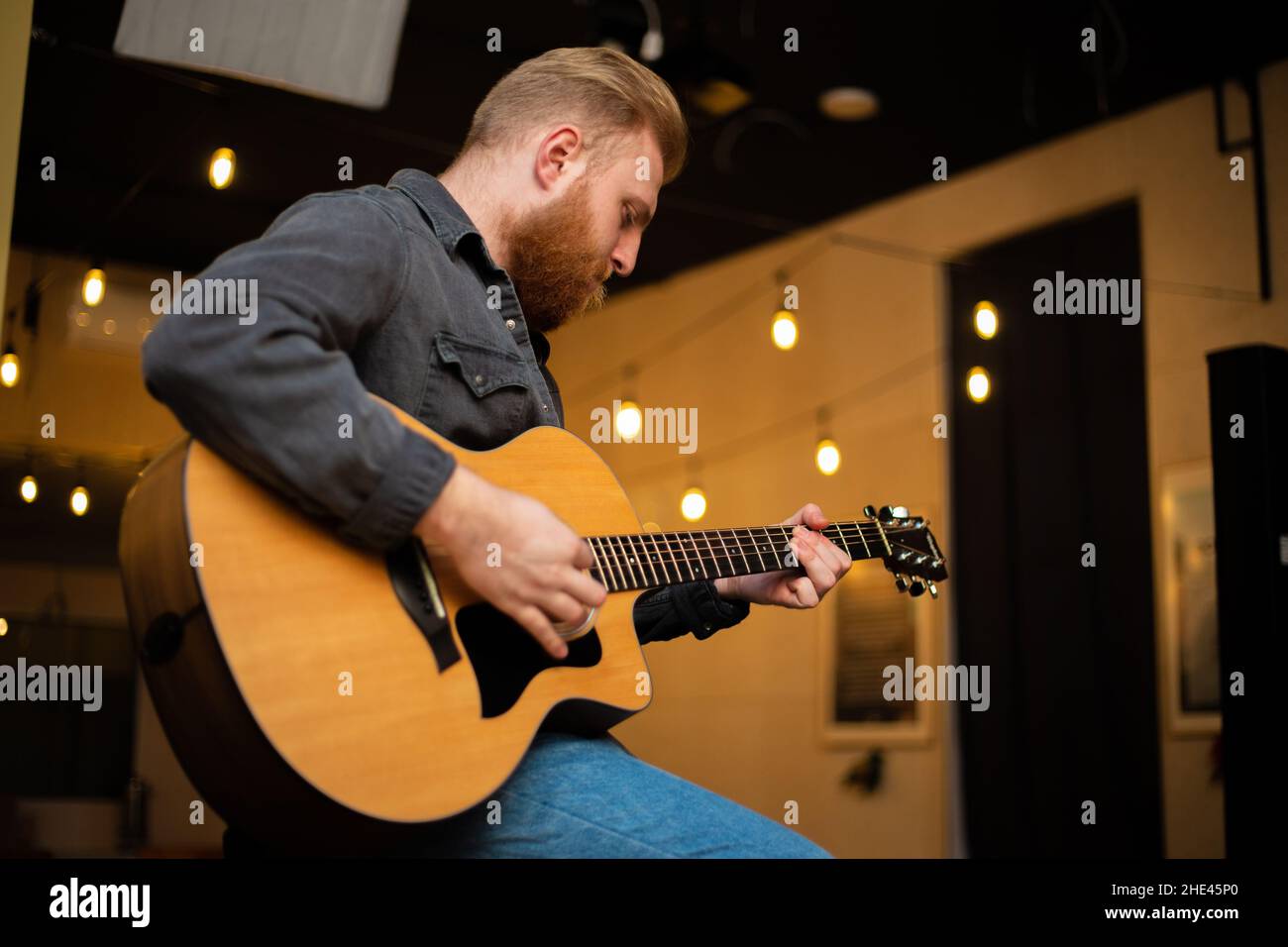 A young guy with a beard plays an acoustic guitar in a room with warm lighting Stock Photo
