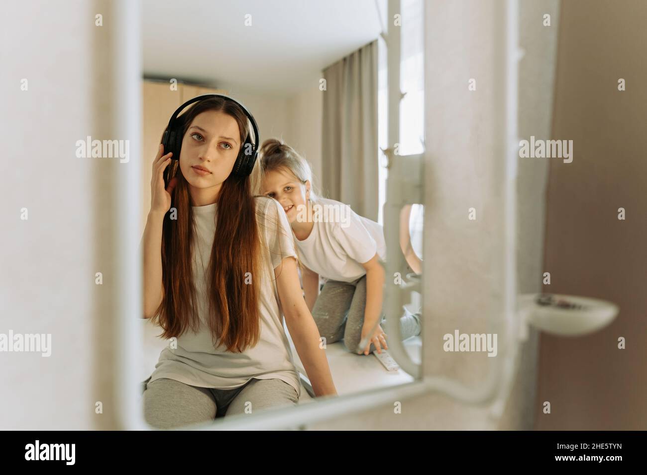 Teenage girl in headphones at mirror, little sister interferes, grimaces. Stock Photo