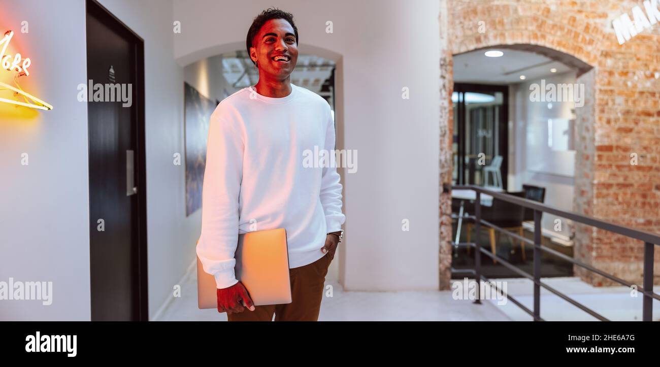 Happy businessman smiling cheerfully while standing alone in a modern workplace. Young businessman holding a laptop in his hand. Creative businessman Stock Photo
