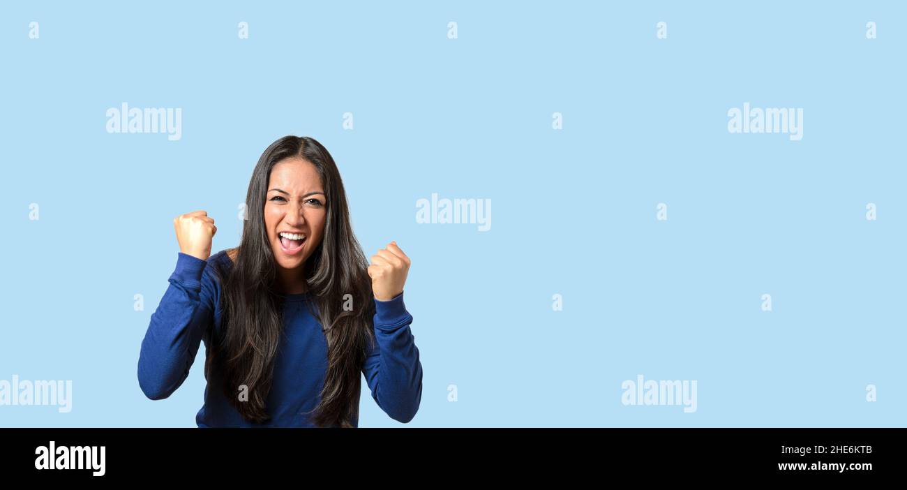 Enthusiastic young woman throwing a temper tantrum balling her fists and yelling at the camera over blue with copyspace Stock Photo