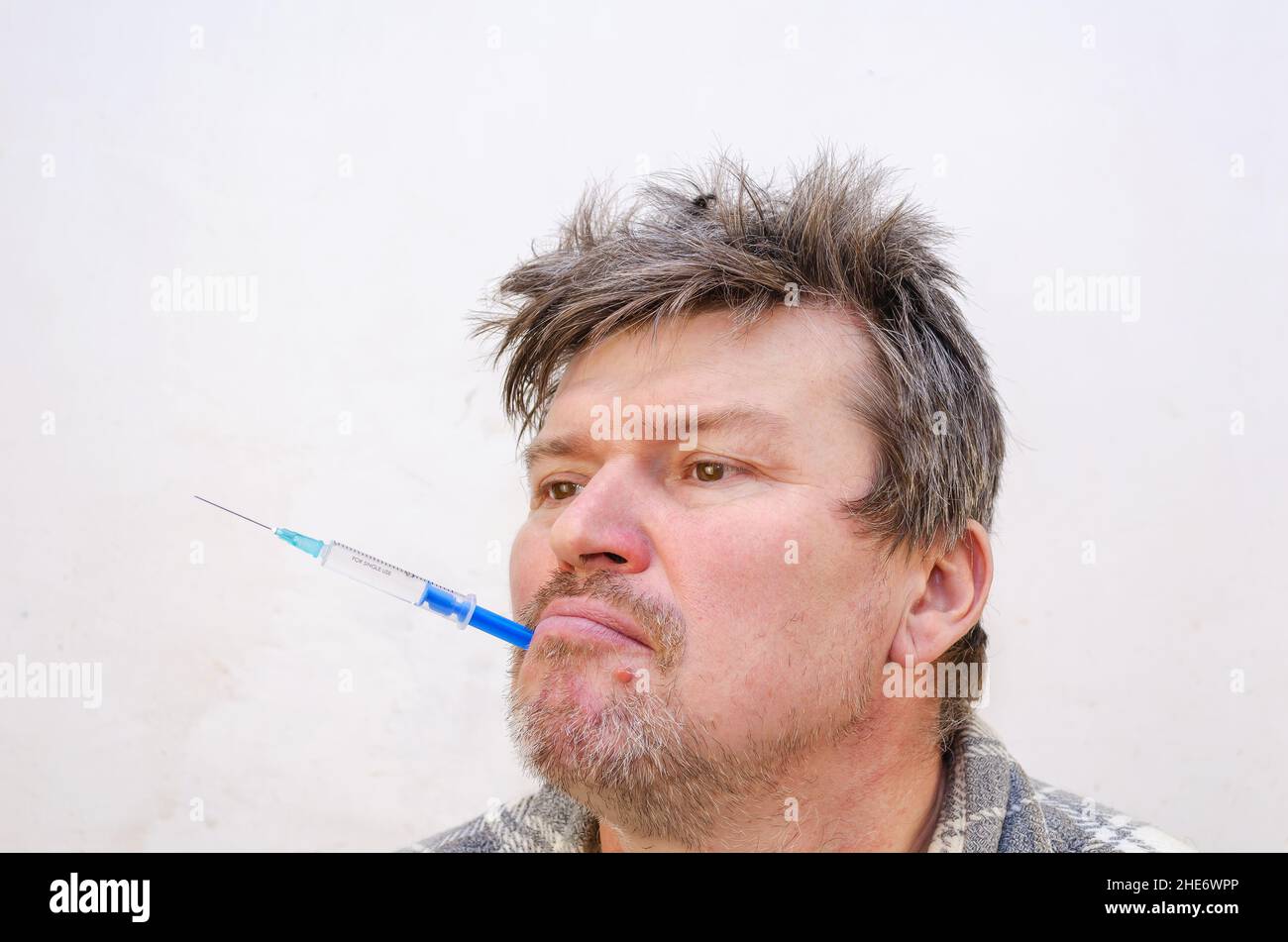 Portrait of a weirdo holding a blue syringe in his mouth. Unshaven man holding a disposable syringe full of liquid. Adult male with ruffled hair.  Ind Stock Photo