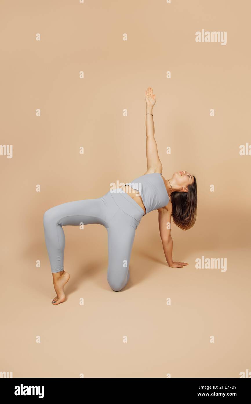 Young brunette is engaged in sports. Beautiful woman in gray tight-fitting yoga suit stands in asana pose, she raised her hand up and stands on one Stock Photo