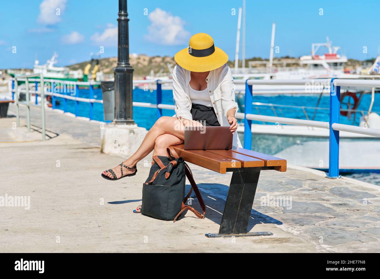 Mature woman in a hat with a laptop on the sea promenade in the city ...