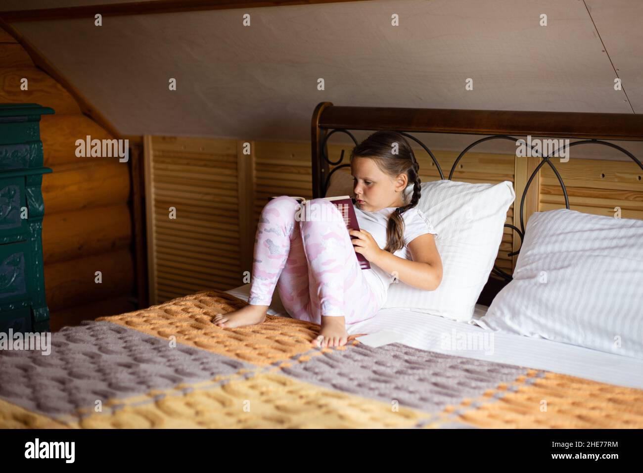 Small keen female kid with book reading with big attention sitting on bed wearing pajamas in wooden house. Free time leisure activities. Self Stock Photo