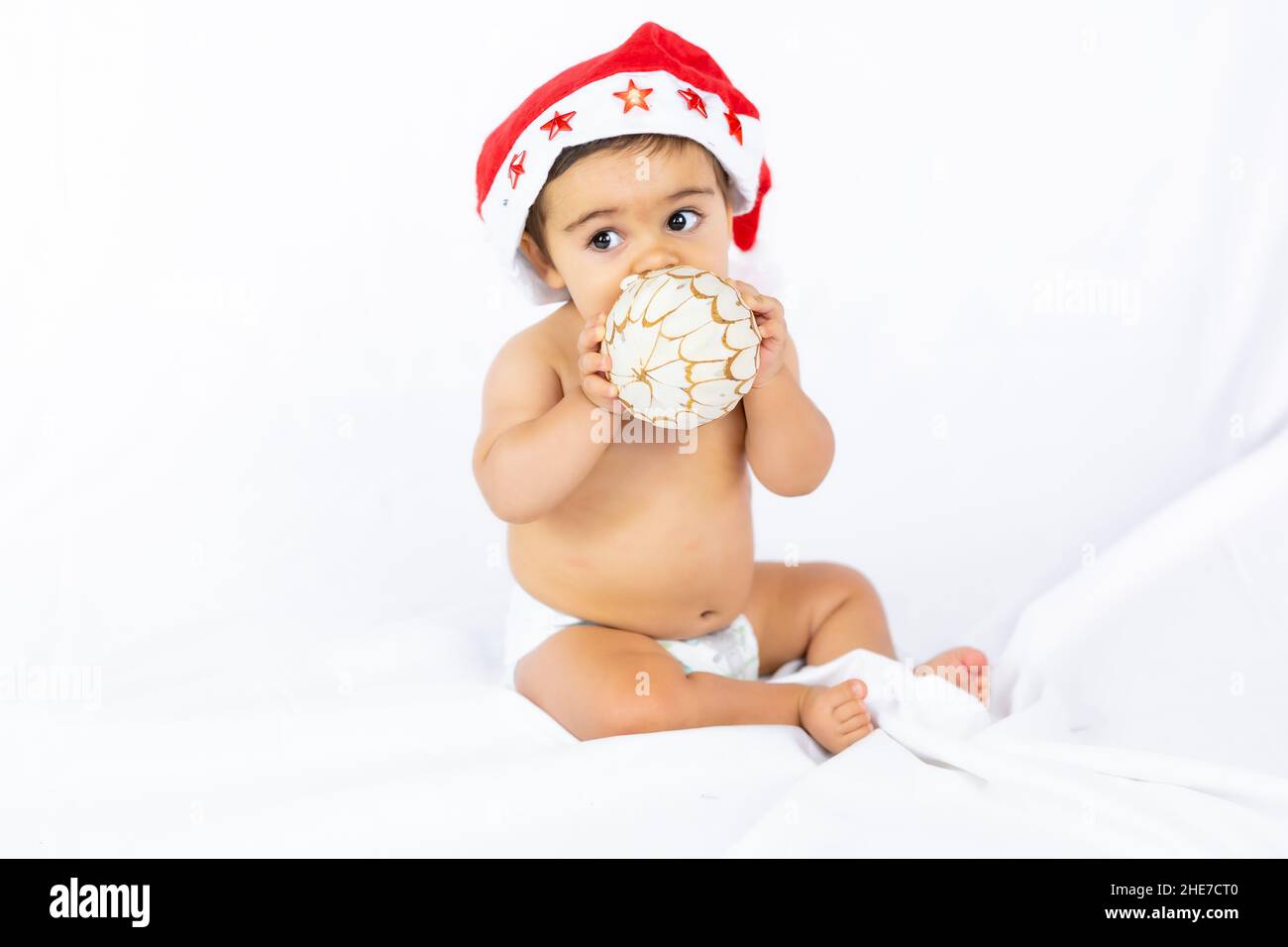 A baby boy with a red Christmas hat on a white background, copy space, with Santa's hat playing with a Christmas tree ball Stock Photo