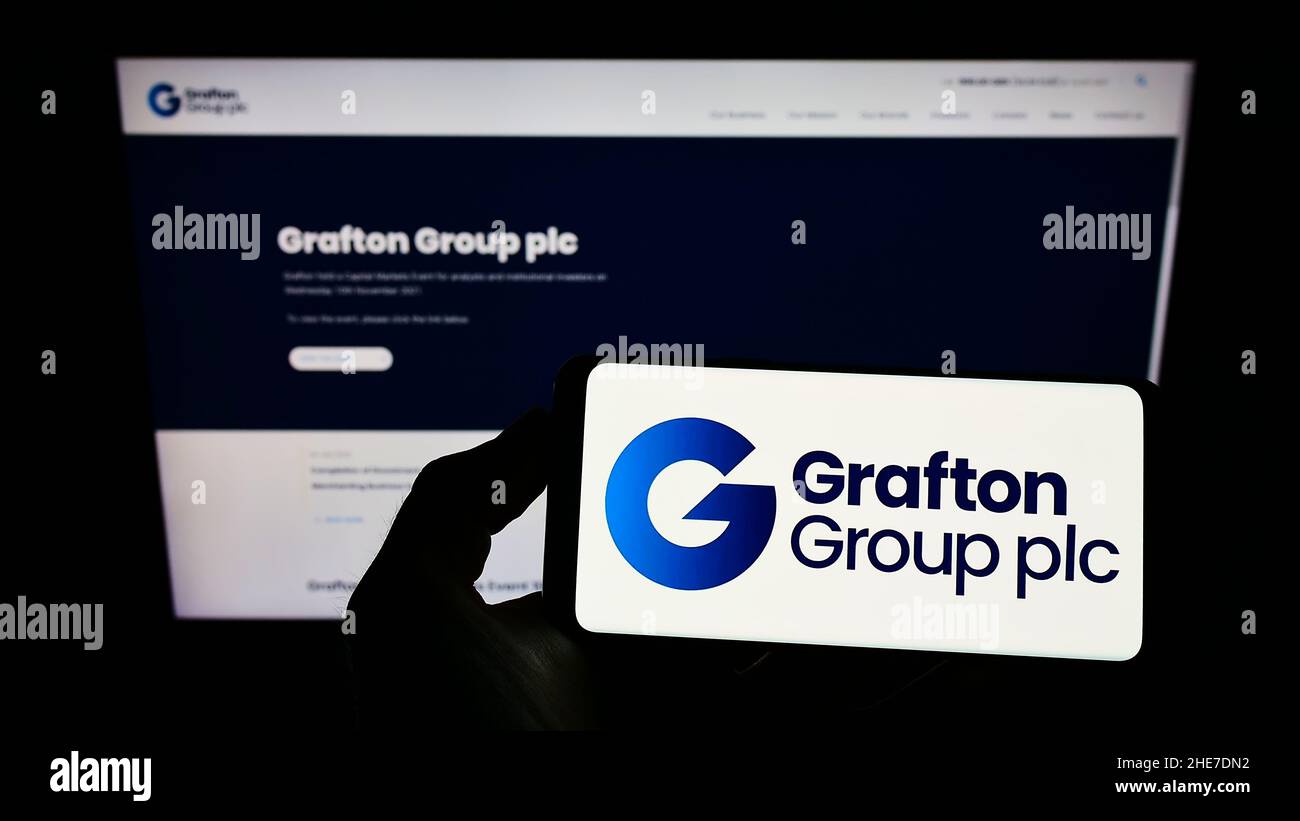 Person holding smartphone with logo of builders merchant company Grafton Group plc on screen in front of website. Focus on phone display. Stock Photo