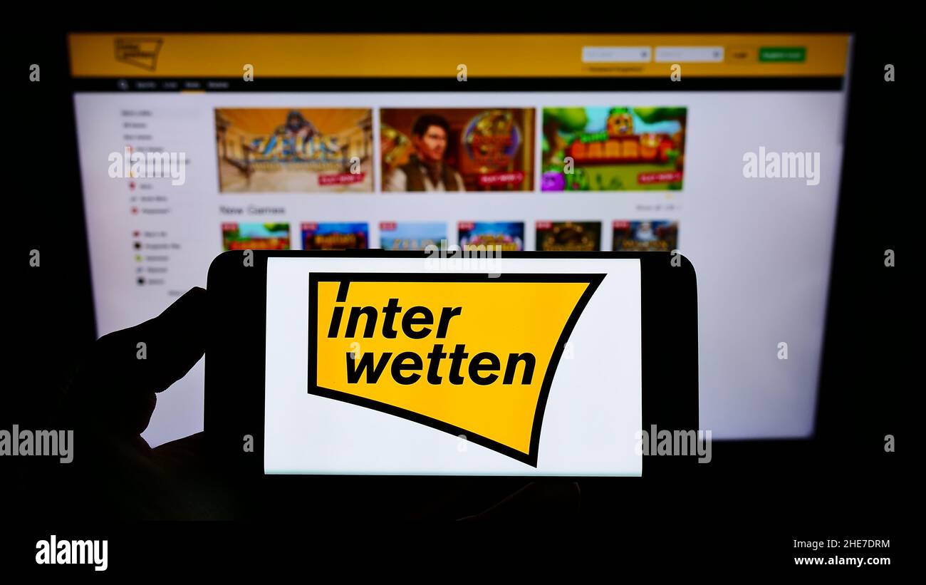 Person holding smartphone with logo of sports betting company Interwetten Ltd. on screen in front of website. Focus on phone display. Stock Photo