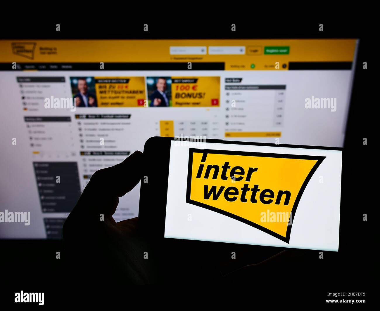 Person holding mobile phone with logo of sports betting company Interwetten Ltd. on screen in front of business web page. Focus on phone display. Stock Photo