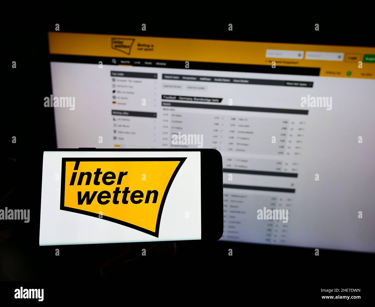 Person holding cellphone with logo of sports betting company Interwetten Ltd. on screen in front of business webpage. Focus on phone display. Stock Photo