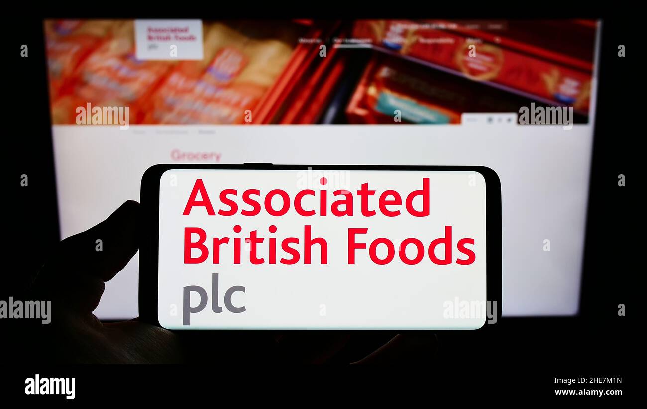 Person holding mobile phone with logo of company Associated British Foods plc (ABF) on screen in front of web page. Focus on phone display. Stock Photo