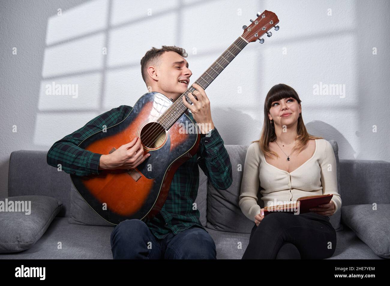 Couple having a great time, young man is playing guitar while the girl is annoyed by loudness, trying to read a book Stock Photo