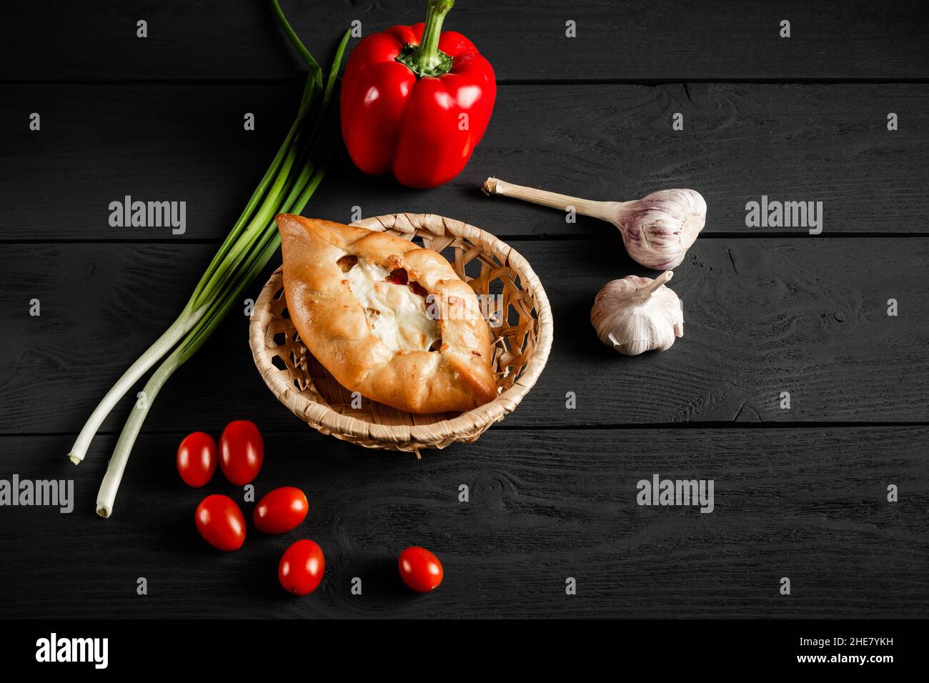 Pie in a wicker basket on a black wooden background. With a copy space.Vegetable composition - tomatoes, pepper, onion, garlic. Stock Photo