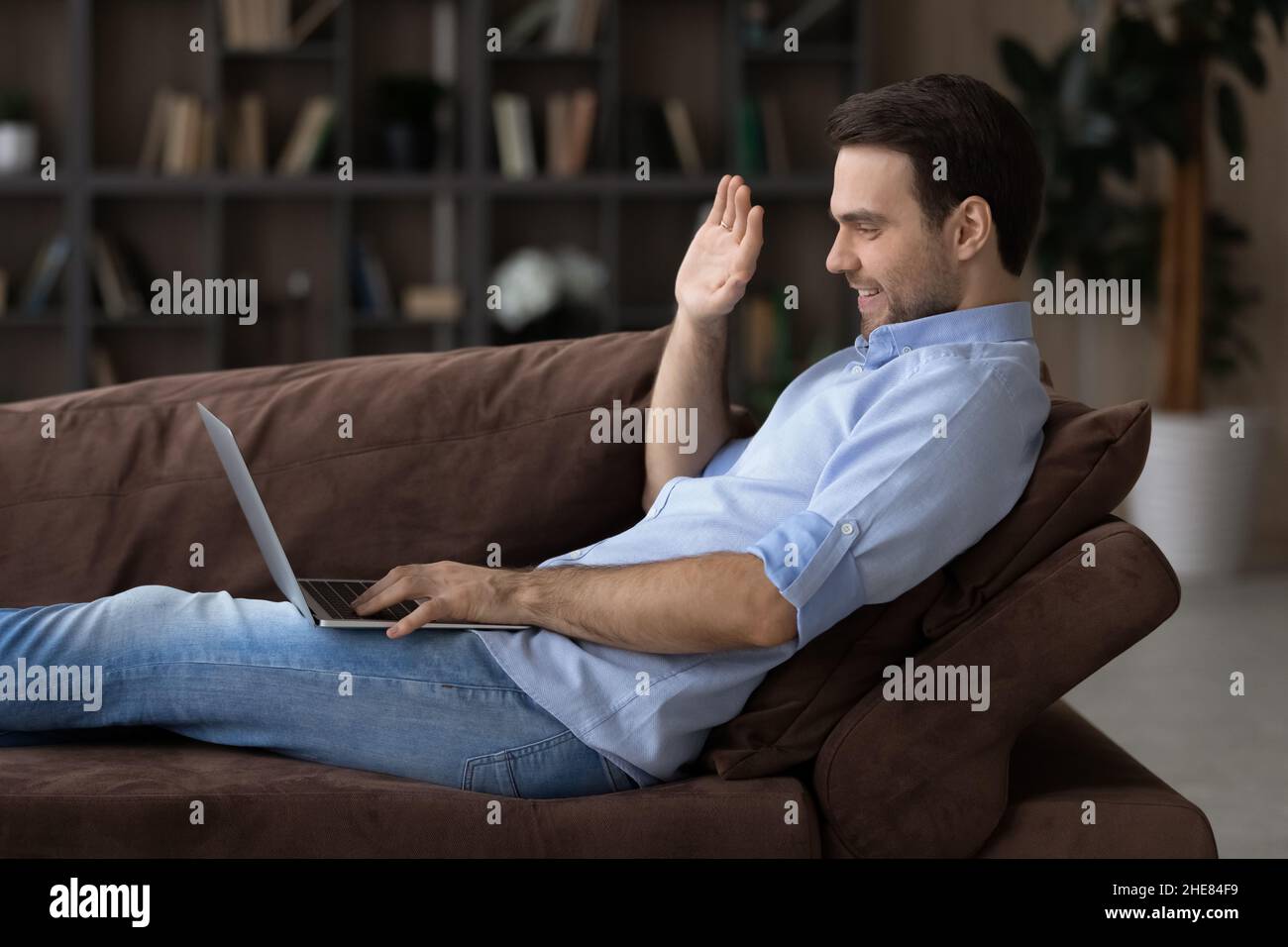 Profile view smiling man waving hand, using laptop on couch Stock Photo