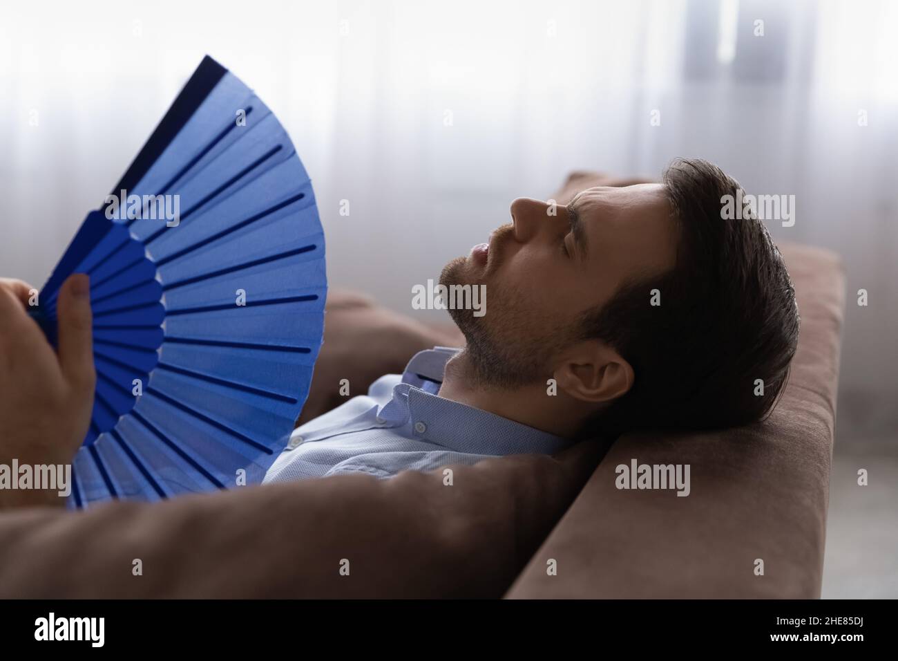 Head shot exhausted man waving paper fan, suffering from overheating Stock Photo