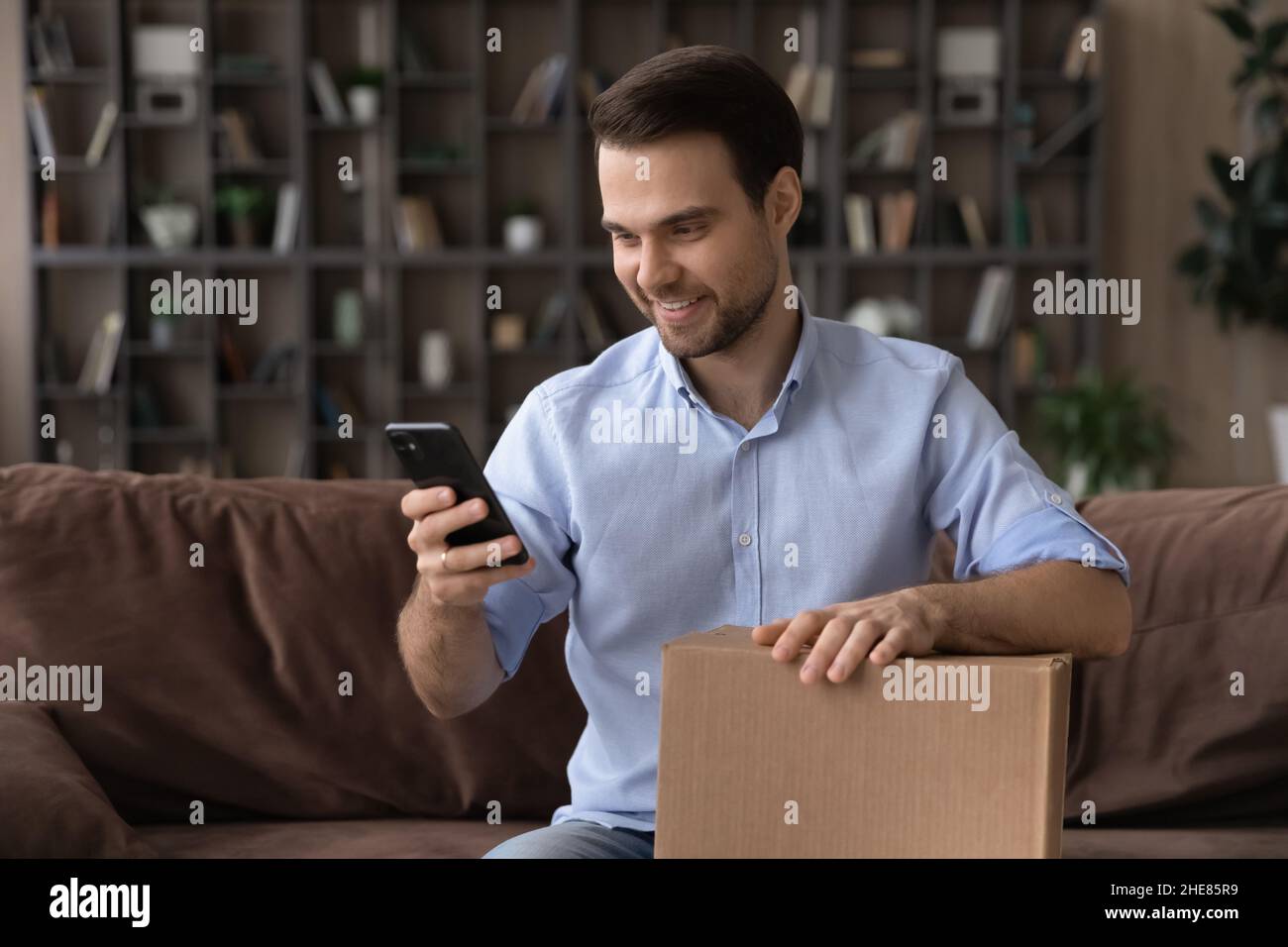 Smiling man using smartphone, holding cardboard box, unpacking parcel Stock Photo