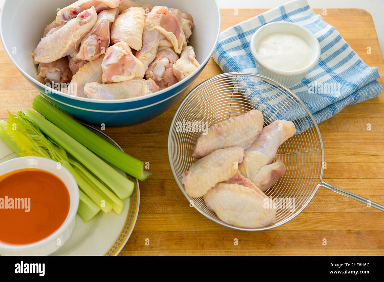 Fresh raw Chicken wings ready for deep frying with a strainer , hot sauce , blue cheese sauce and celery on a wooden cutting board in a home kitchen Stock Photo