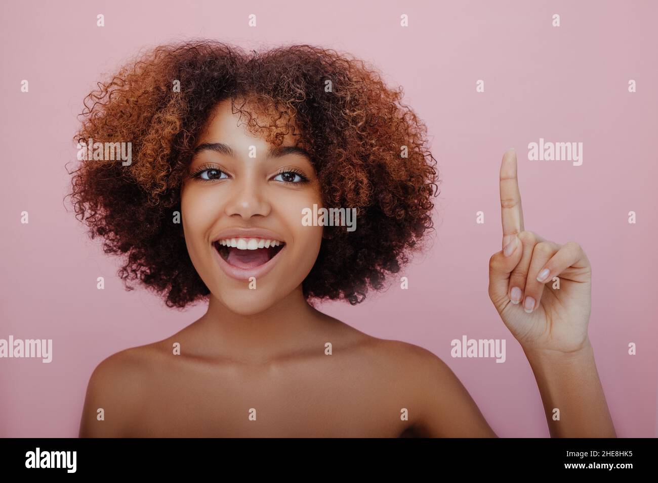 Young beautiful African-American woman with a lush curly hairstyle points her index finger up looks at the camera and smiles on a pink background, a new idea has come, inspiration Stock Photo