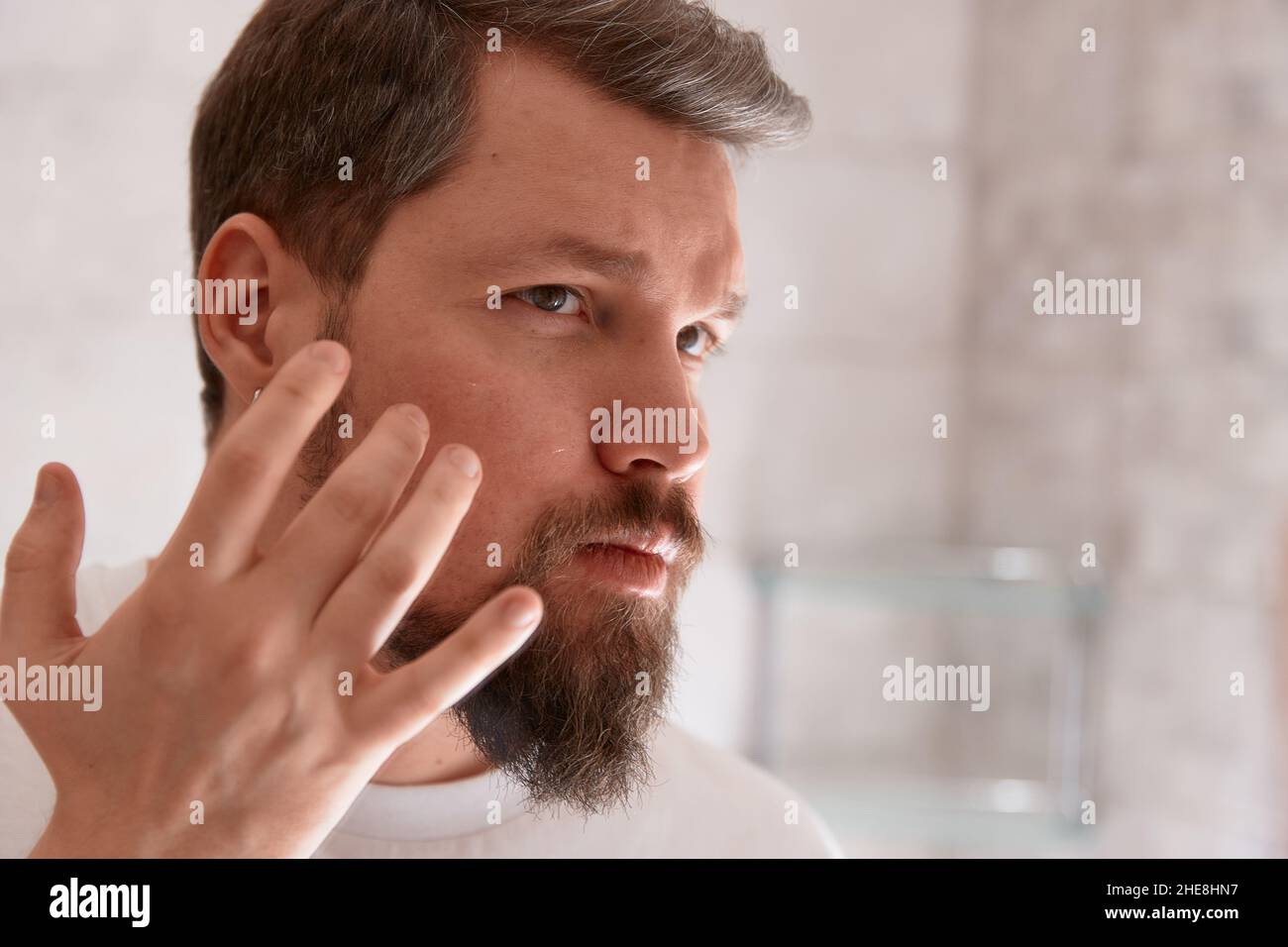Portrait of man in white tshirt applying serum on his face at bathroom. Self care morning procedure Stock Photo